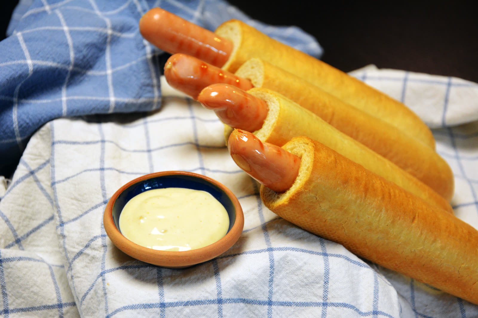 Fire ferdige franske hotdoger med pølse i hullet ligger på et blårutet kjøkkenhåndkle ved siden av en liten skål med lys dressing