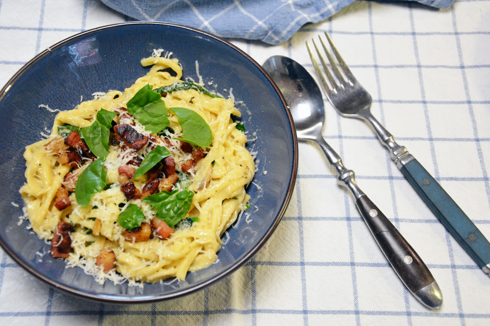 Kremet pasta med bacon, spinat og revet parmesan i en blå skål på et blårutet kjøkkenhåndkle
