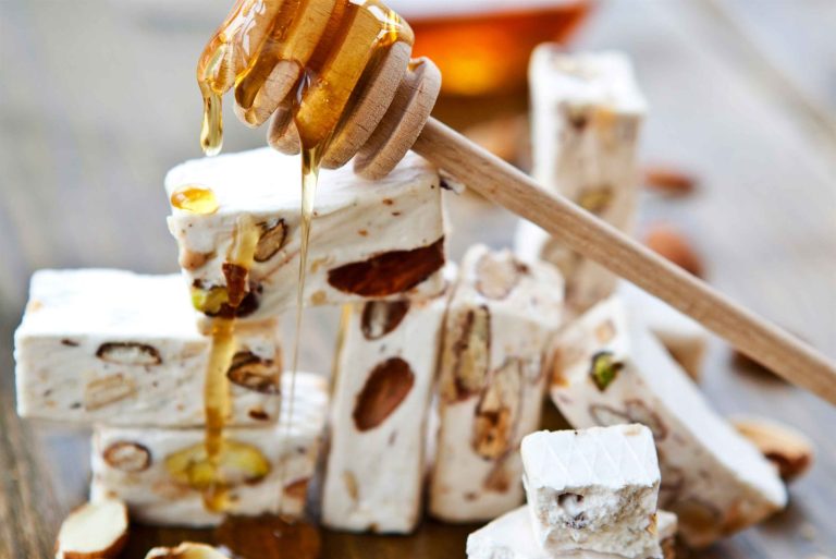 Biter av hvit fransk nougat med hele mandler og pistasjnøtter stablet på et trebord mens honning drypper ned over dem