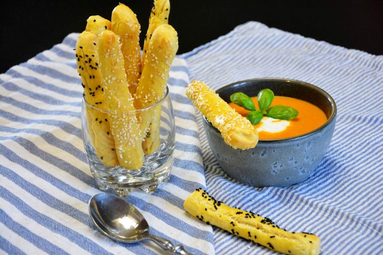 Hjemmelagde grissini med sesamfrø og svartkarve i et glass ved siden av en skål tomatsuppe