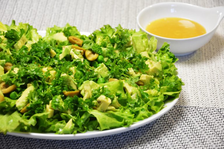 Grønn salat med kruset salat, avokado, grønne oliven, erter og persille på et hvitt fat, med vinaigretten i en liten skål ved siden av
