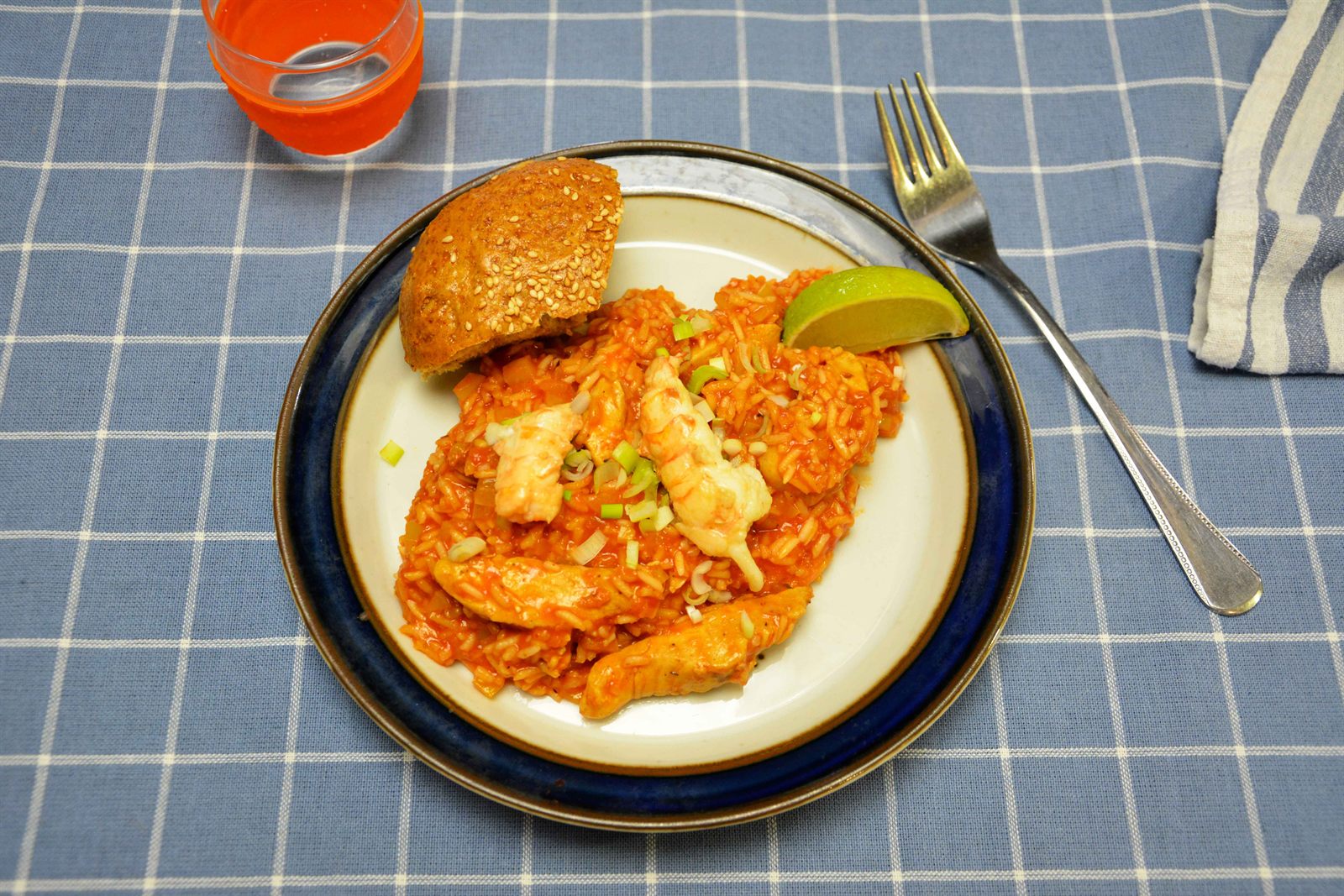 Jambalaya med rød tomatris, kyllingstrimler og sjøkrepshaler servert på en hvit tallerken med en limebåt, hakket vårløk og et sesambrød