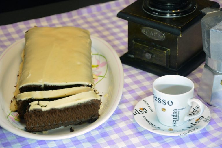 Kaffekake med lys kaffeglasur, skåret i skiver og lagt på et hvitt fat, med espressokopp, gammel kaffekvern og mokkakanne ved siden av