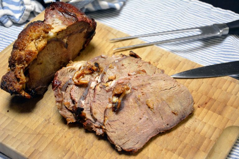 En grillet kalvefilet ligger på et lyst skjærebrett i bambus, med en sprø, gyllenbrun, rutet fettkappe på toppen. Halvparten er skåret i tykke skiver med en lys, lett rosa-grå midte. En forskjærgaffel og en forskjærkniv ligger ved siden av, og et blå-hvitstripet kjøkkenhåndkle ligger i bakgrunnen. Ingen grill er synlig, og det er ikke noe tilbehør på bildet.