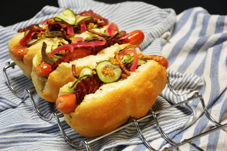 Pølse i brød med bacon, syltet rødløk, sprøstekt løk, agurksalat og syltede sennepsfrø