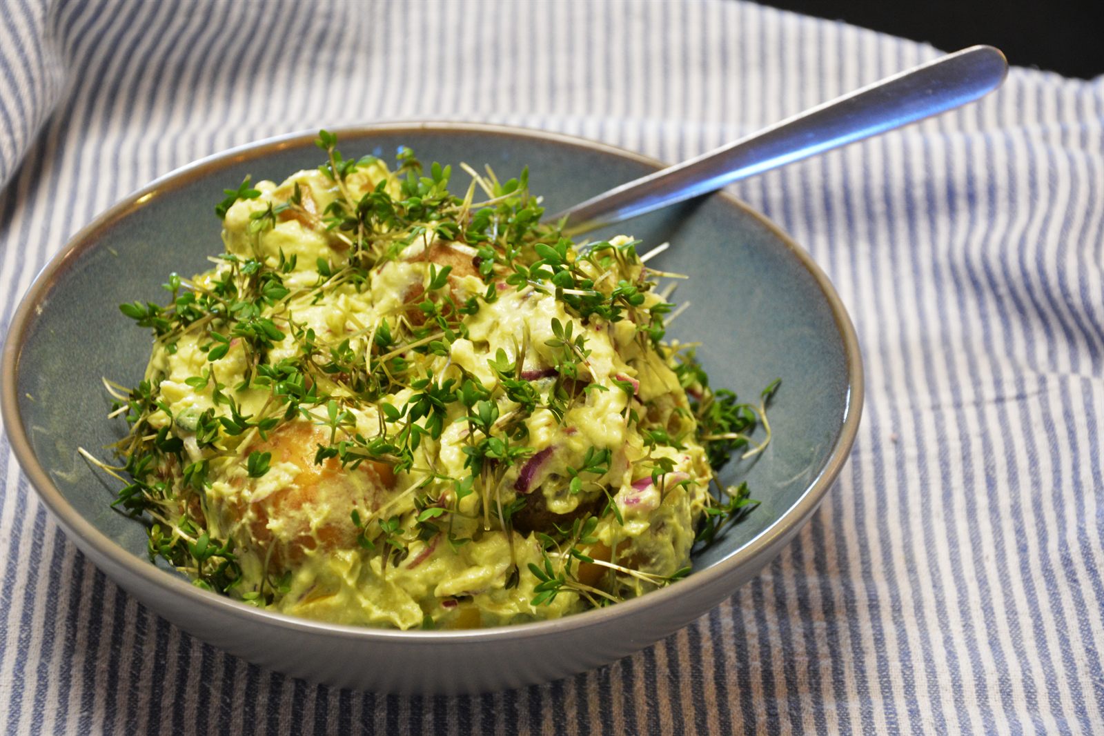 Grønn potetsalat med avokado, kremost og rødløk i en blå bolle, dekket av nyklippet karse