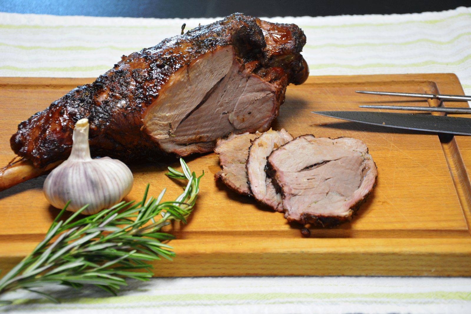 Grillet lammelår med bein på et skjærebrett i tre, som ligger på en stripete linduk. Beinet stikker ut til venstre, overflaten er mørk, nesten svart av grill og røyk, og et snitt i kjøttet viser rosa innside. Tre skiver ligger foran til høyre, et helt hvitløkshode og rosmarinkvister ligger i forgrunnen til venstre, og en kniv og en forskjærgaffel ligger til høyre.