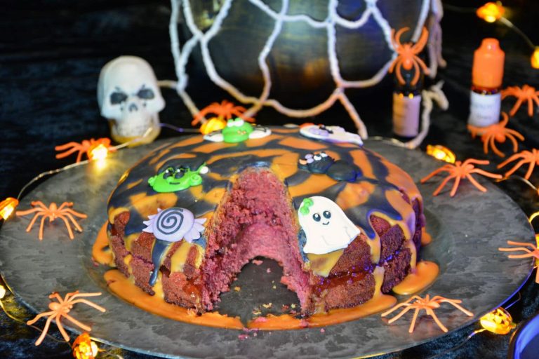 Blodrød halloweenkake med bringebærsyltetøy, rennende oransje og sort glasur og sukkerfigurer, med en bit tatt ut – oppskrift på godtnoe.no