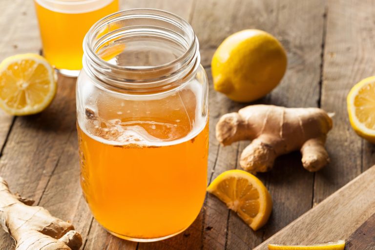 Et åpent glass uten lokk fylt med gyllen, ravfarget kombucha med litt skum på overflaten, på et rustikt trebord; rundt glasset ligger friske ingefærrøtter, hele og halve sitroner og en sitronbåt; et glass kombucha står i bakgrunnen.