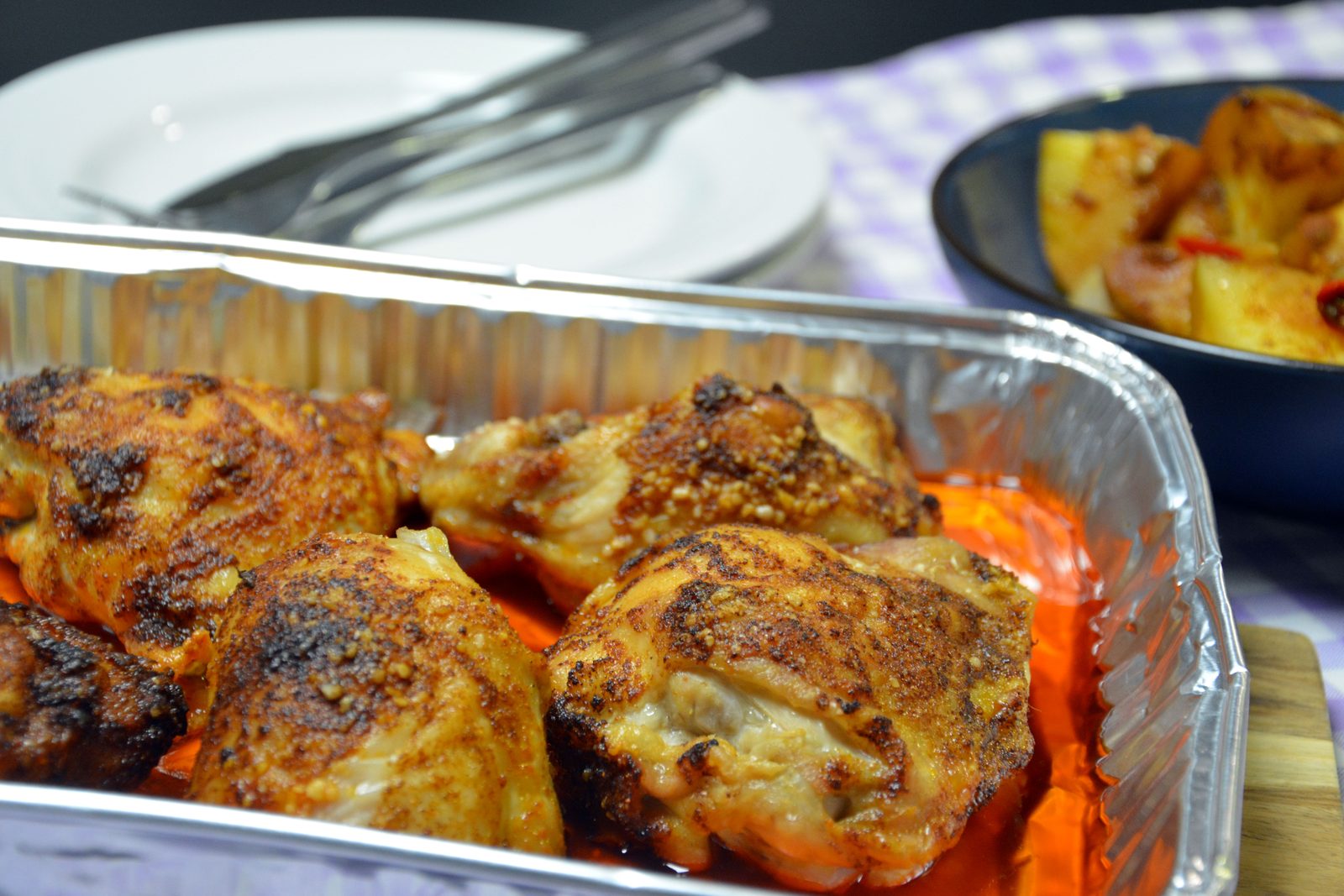 Fire gyldenbrune, paprikakrydrede kyllingoverlår med skinn i en rektangulær aluminiumsbakke, med en rødoransje marinade i bunnen. Bakenfor står en hvit tallerken med kniv og gaffel, og øverst til høyre en mørkeblå skål med ovnsbakte potetbiter og et stykke rød chili. Duken er lilla- og hvitrutet, og bakgrunnen er mørk.