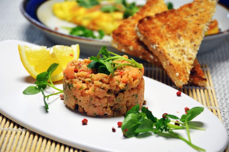 Et rundt tårn av laksetartar med dill og rosa pepperkorn, toppet med brønnkarse, på en avlang hvit tallerken med en sitronbåt og løse rosa pepperkorn. Bak tallerkenen ligger trekanter av ristet kornbrød og en tallerken med omelett, og den avlange tallerkenen står på en bambusmatte.