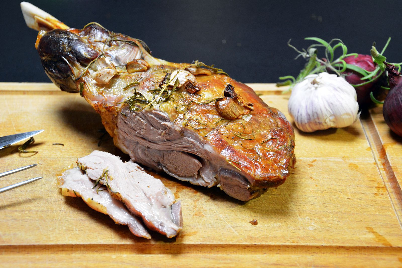 Ovnsstekt lammebog med bein på et skjærebrett i tre, med sprø, brun overflate, bakte hvitløksfedd og rosmarinnåler. Knokkelen stikker opp til venstre, to skiver rosa kjøtt ligger foran, en kniv og en gaffel ligger til venstre, og et helt hvitløkshode, rødløk og rosmarinkvister ligger til høyre mot en mørk bakgrunn.