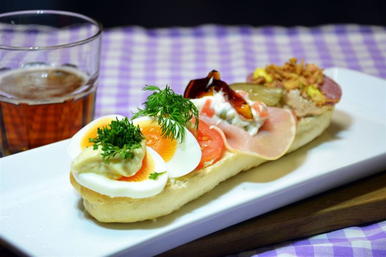Et landgangsbrød, en halv baguette med åpen overside, på et langt, rektangulært hvitt fat. Fremst ligger kokte eggbåter med en klatt majones og karse, så tomatskiver med en dillkvist, så en skive hamburgerrygg med italiensk salat av erter og gulrot og et stykke kjøttgelé, og bakerst leverpostei og spekepølse med stekt løk og en skive syltet agurk. Til venstre står et ølglass. Duken er lilla- og hvitrutet, og bakgrunnen er mørk.