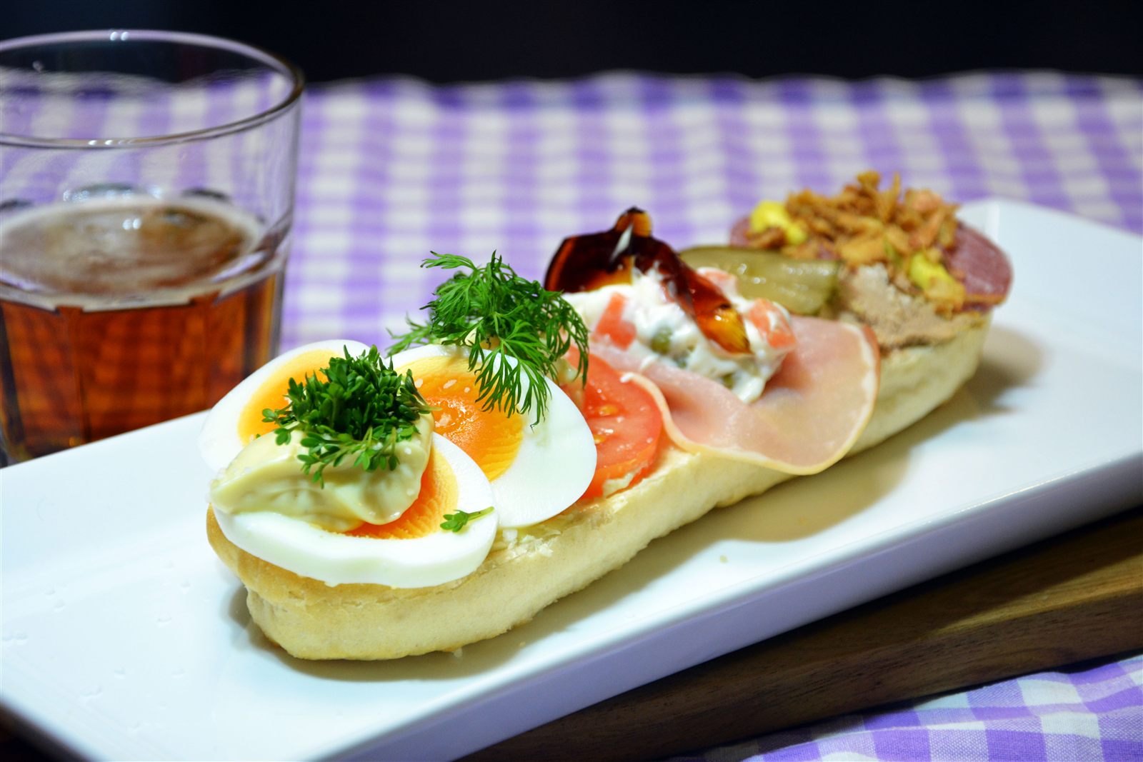 Et landgangsbrød, en halv baguette med åpen overside, på et langt, rektangulært hvitt fat. Fremst ligger kokte eggbåter med en klatt majones og karse, så tomatskiver med en dillkvist, så en skive hamburgerrygg med italiensk salat av erter og gulrot og et stykke kjøttgelé, og bakerst leverpostei og spekepølse med stekt løk og en skive syltet agurk. Til venstre står et ølglass. Duken er lilla- og hvitrutet, og bakgrunnen er mørk.
