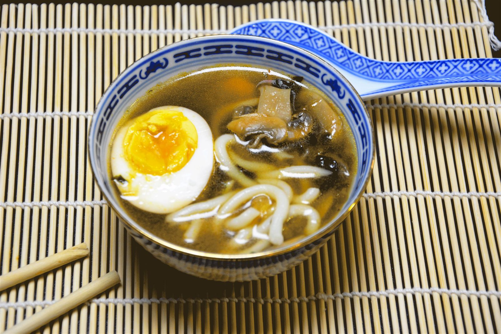 En blå og hvit porselensskål med mørk, klar suppe, tykke hvite udon-nudler, sjampinjongbiter og et halvt egg med gul plomme. Bak skålen ligger en blå og hvit porselensskje, foran ligger to spisepinner av tre, og alt står på en lys bambusmatte.