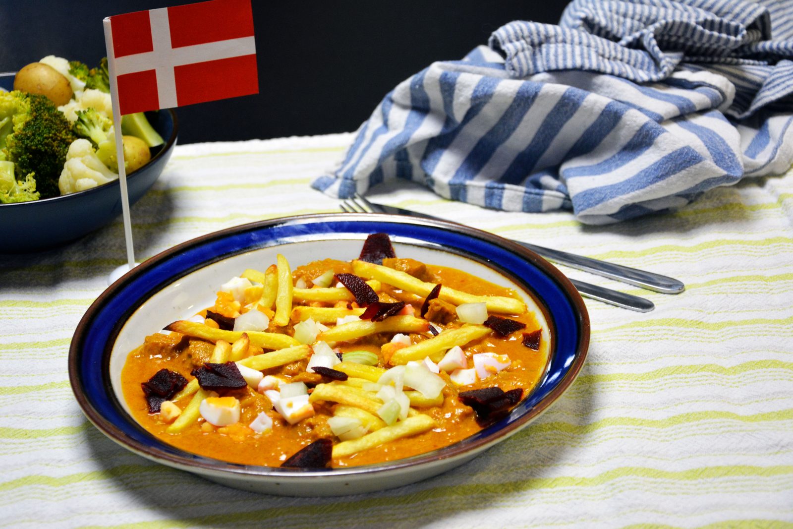 En dyp tallerken med blå kant, fylt med kjøttstykker i oransje fløtesaus, toppet med pommes frites, hakkede hardkokte egg, hakket rå løk og biter av syltede rødbeter. Til venstre står et lite dansk flagg (rødt med hvitt kors) og en mørk skål med små kokte poteter, brokkoli og blomkål. Gaffel og kniv ligger ved siden av, med et blåstripet kjøkkenhåndkle og en hvit- og gulstripet duk.